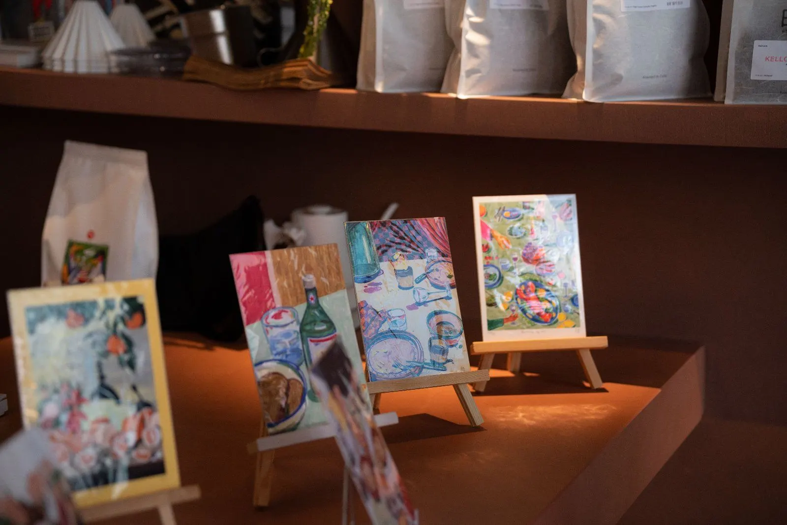 Small colourful paintings stand on easels on a wooden shelf lit by a warm lamp.