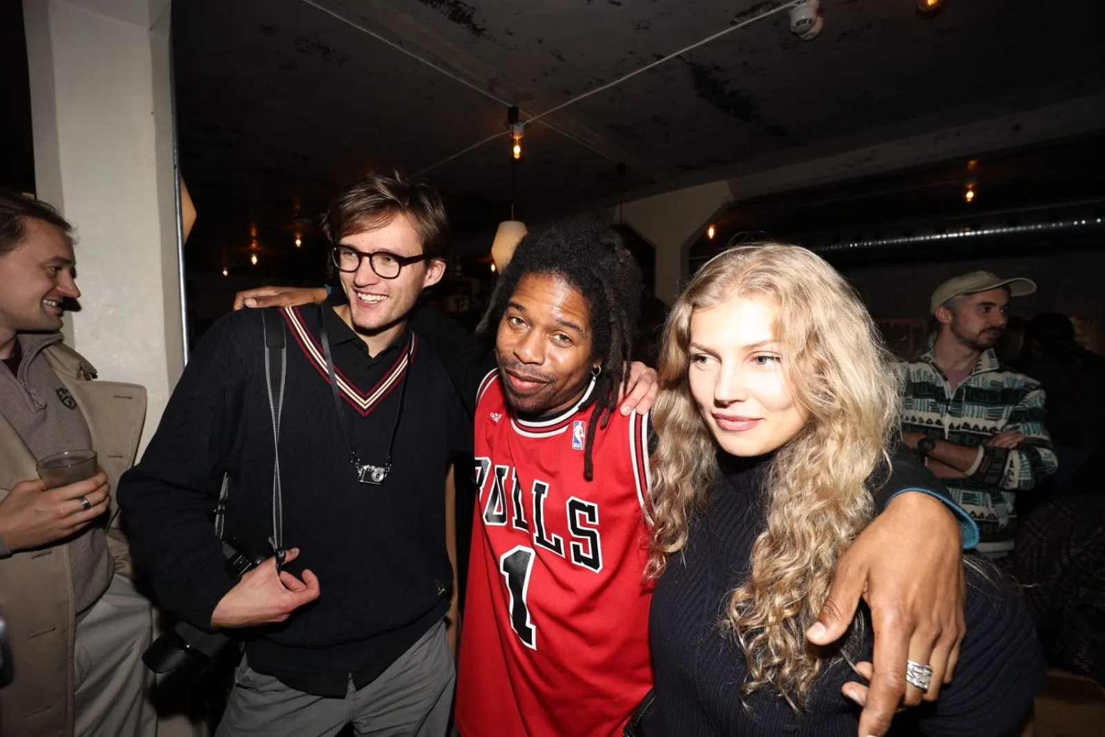 Three friends pose with their arms around each other in the dim bar, one in a red basketball jersey.