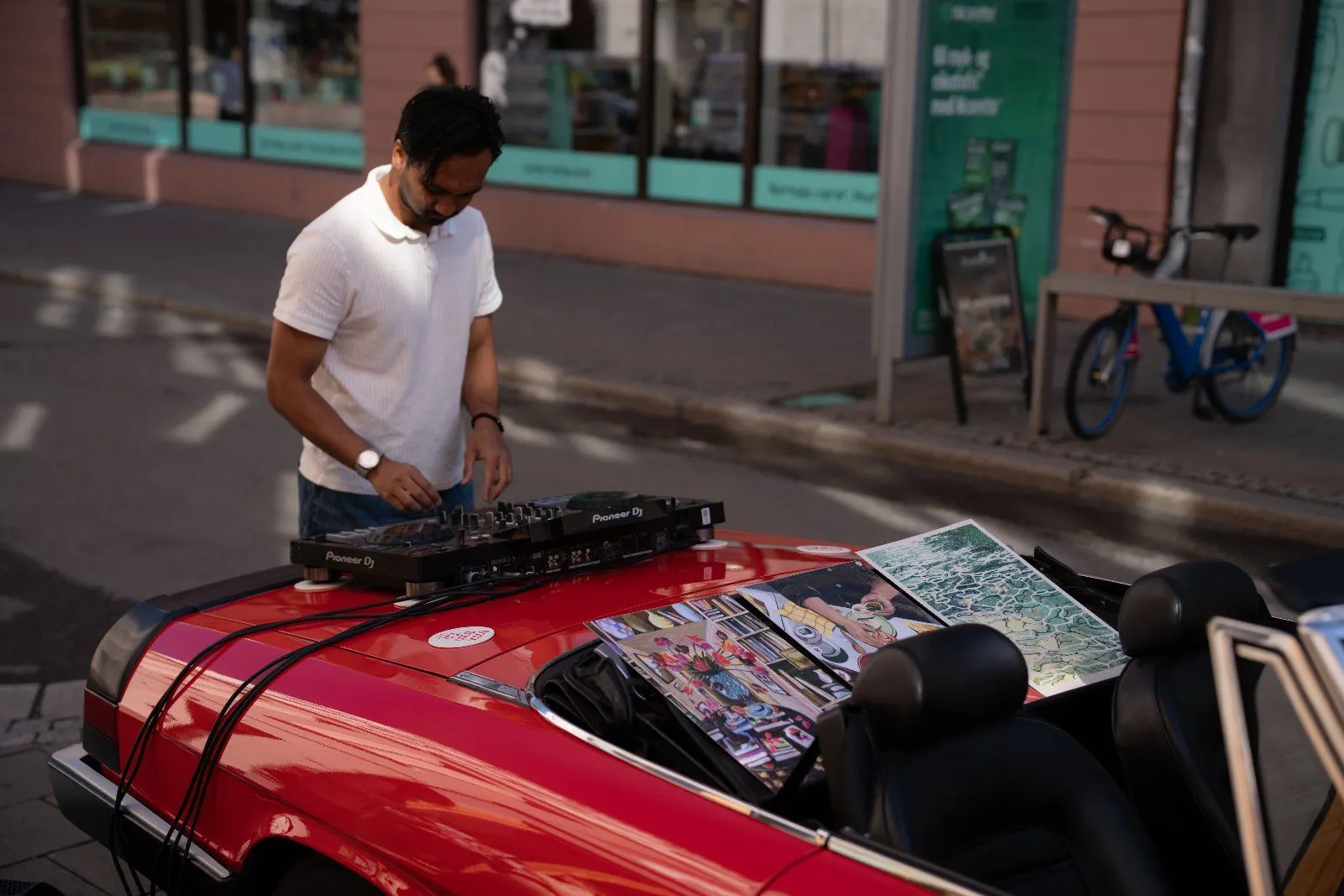 A DJ plays on decks set up on the bonnet of the red convertible, with art prints laid out beside him.