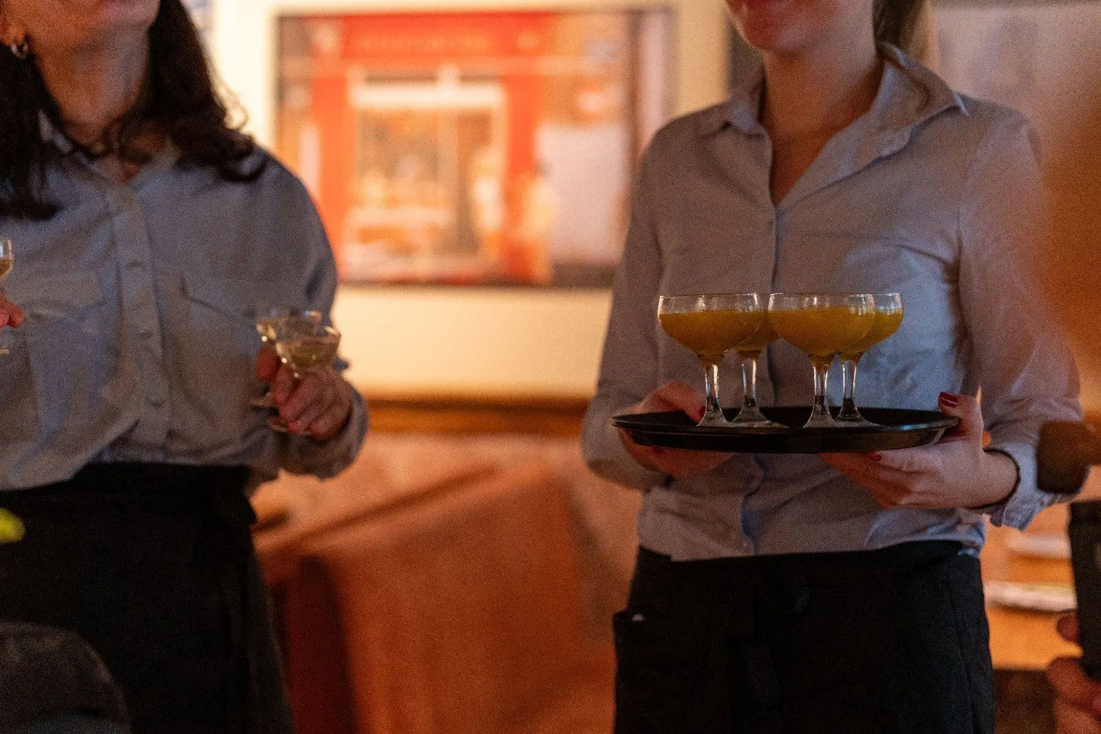Two servers in blue shirts carry a tray of golden cocktails through the room.