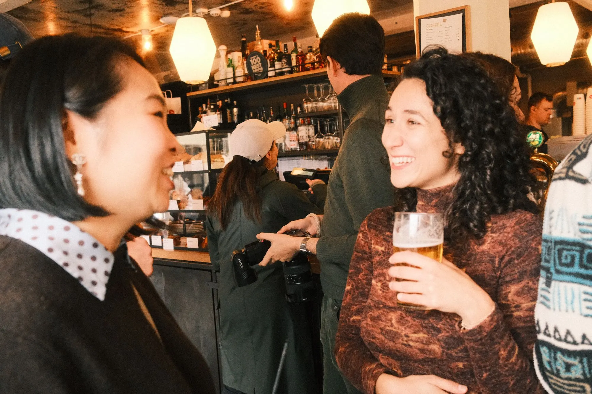 Two women laugh together at the bar under warm hanging lights.