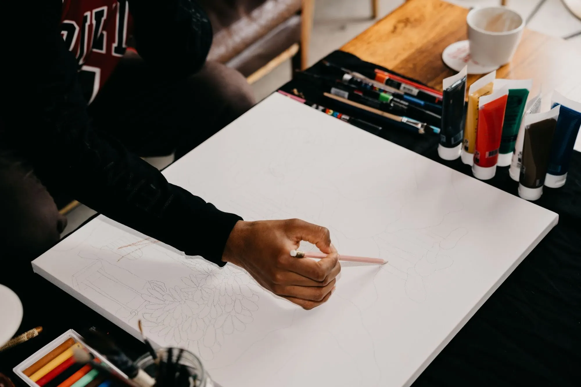A hand sketches on a white canvas with paints and pencils spread across the table.