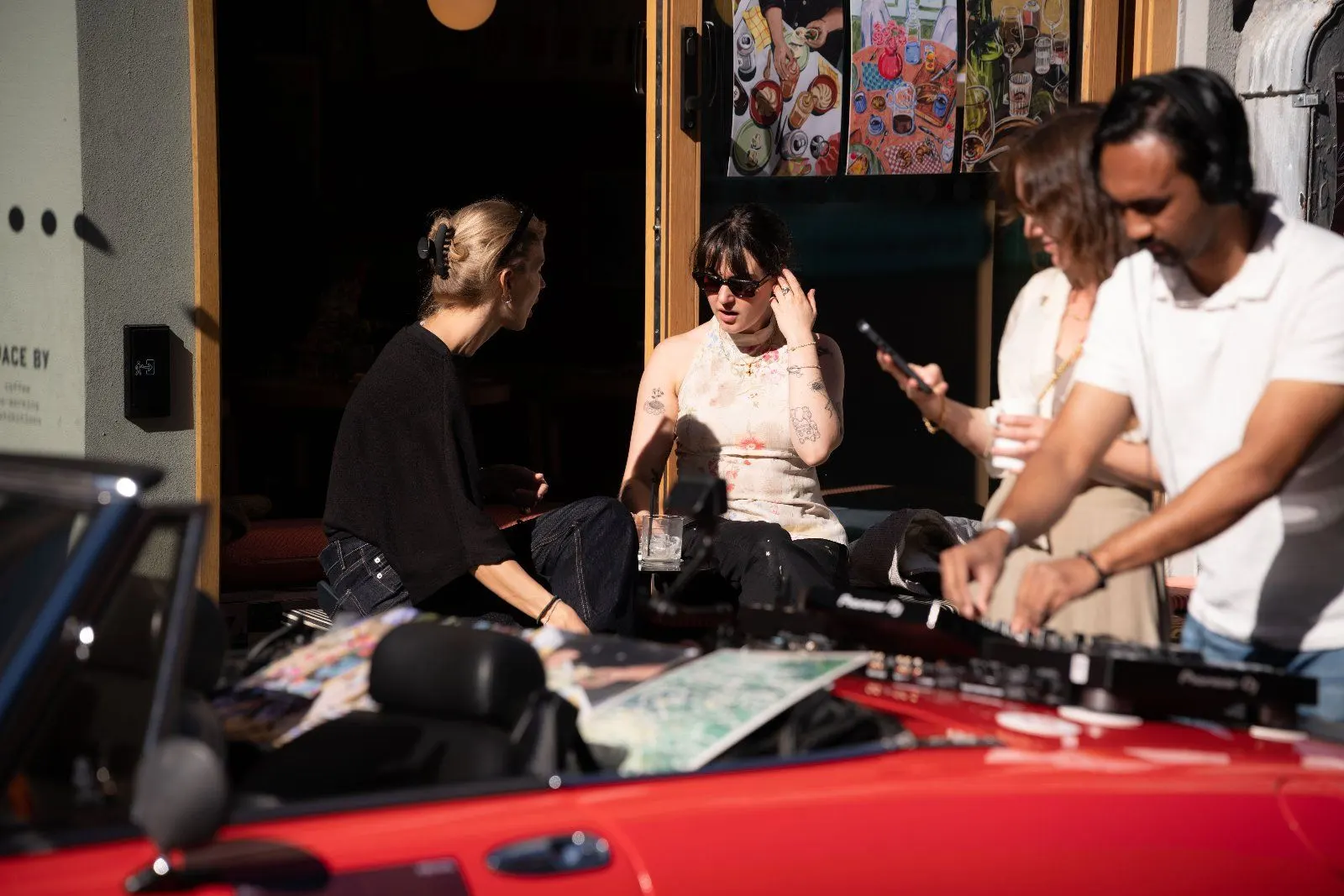 A DJ and guests chat in the open doorway beside the car in strong afternoon sun.
