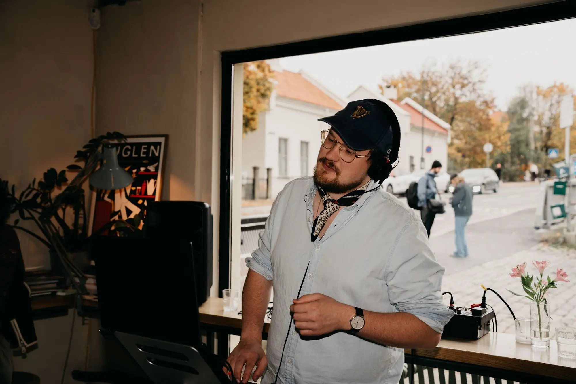A DJ in a light shirt and cap stands by the large window at Fuglen with the autumn street behind him.