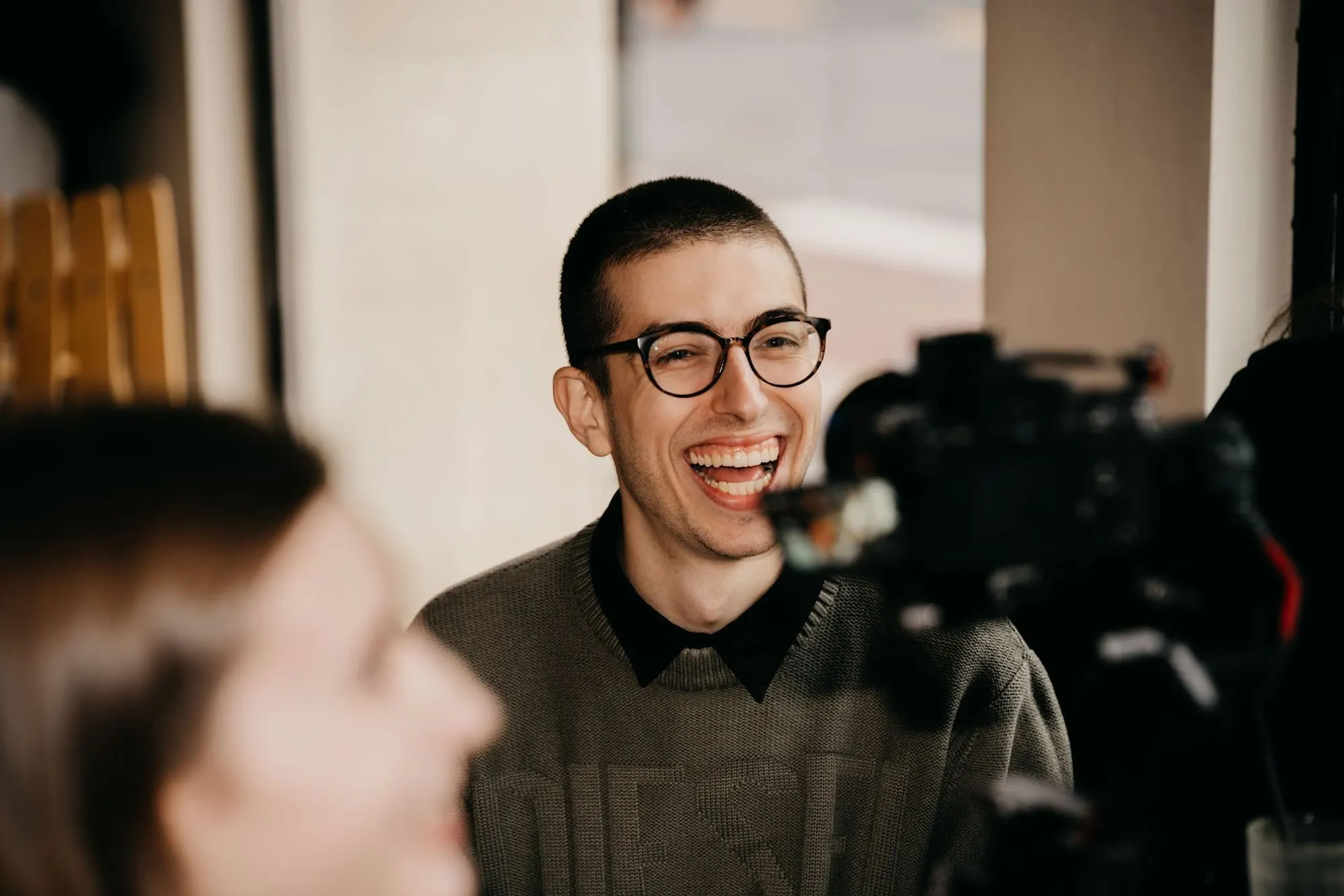 A smiling man with round glasses laughs, with a camera in the foreground.