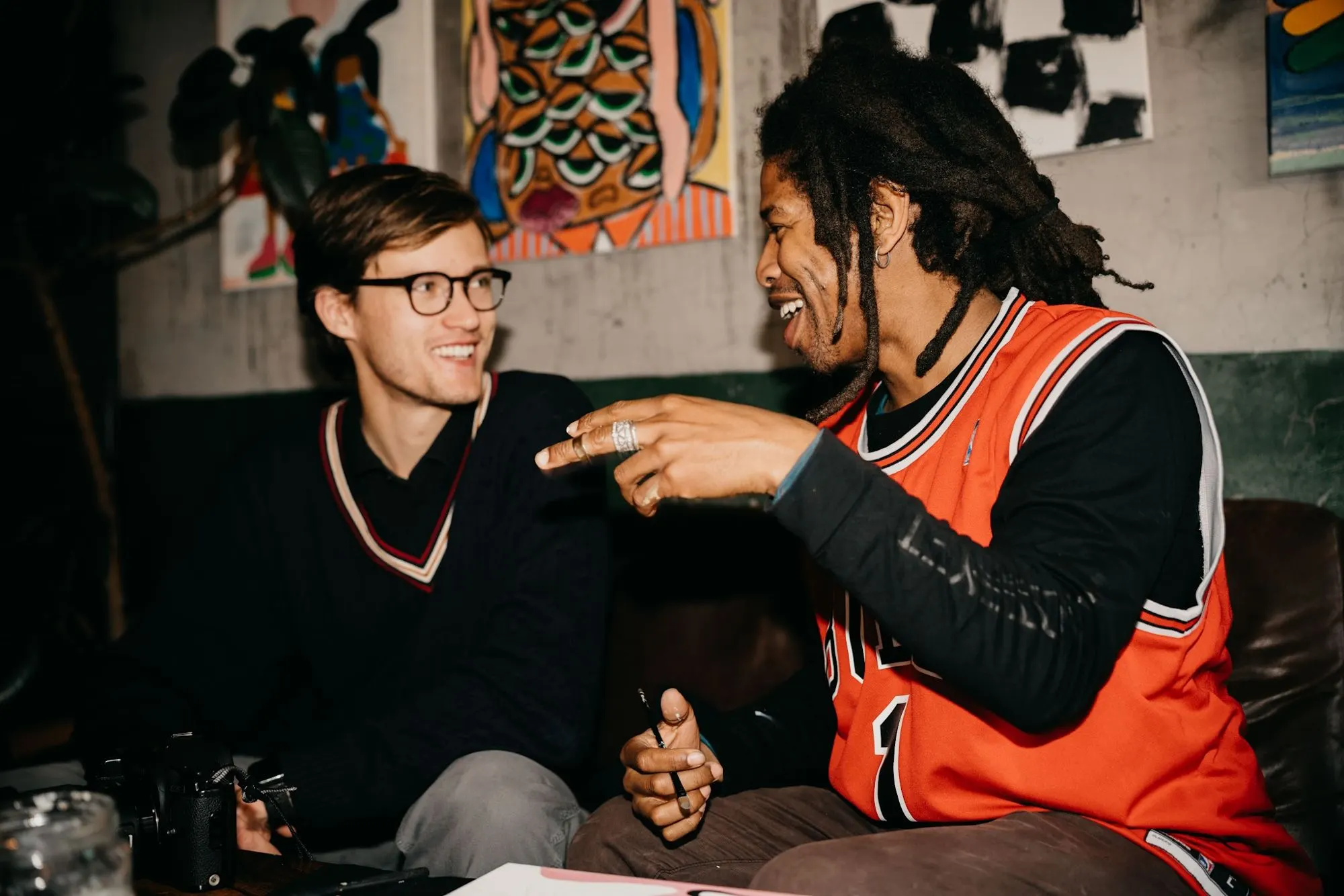 Two men sit on a sofa talking, one in an orange basketball jersey, with colourful art on the wall behind.
