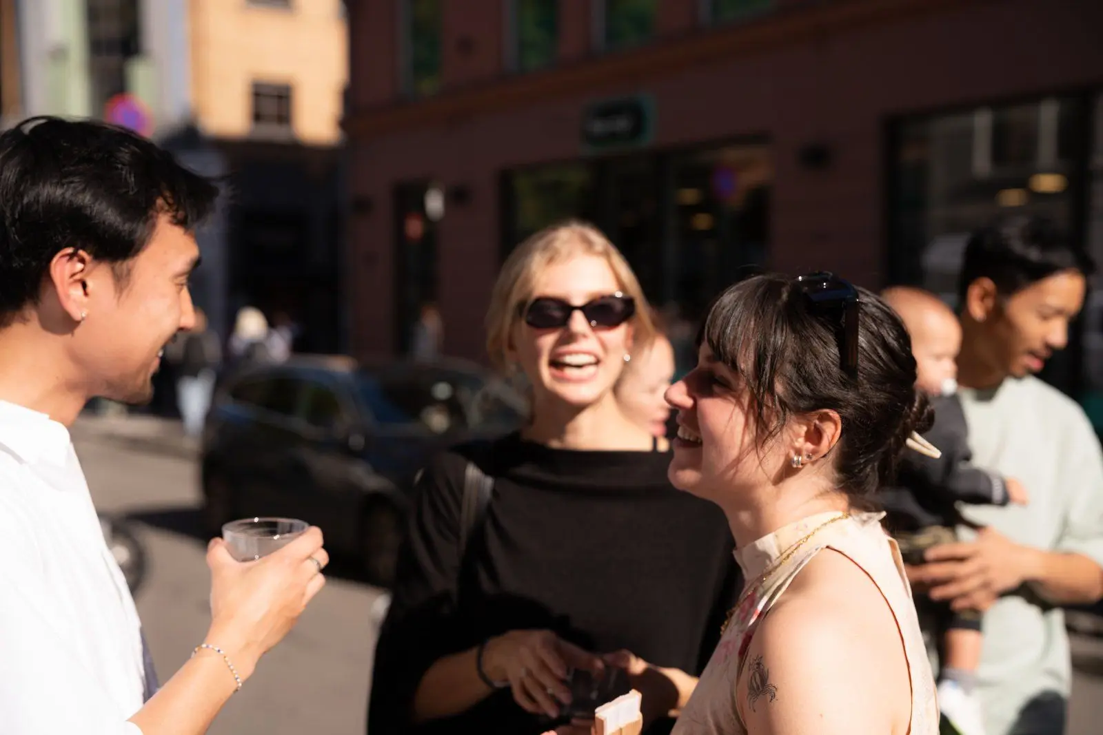 Three guests laugh together on the pavement, one holding a glass.