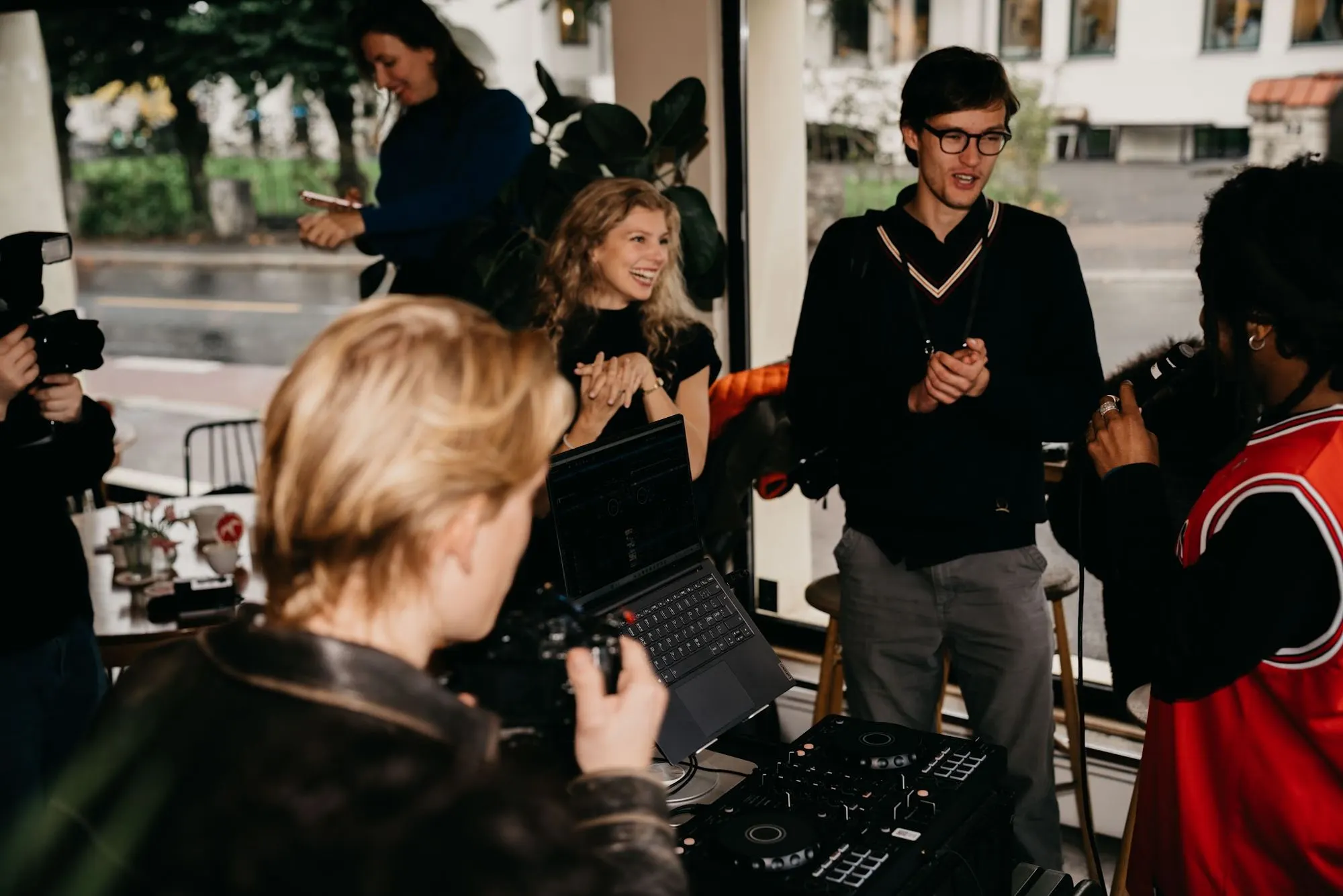Guests gather around the DJ decks by the window while a man in a knit vest talks to the room.