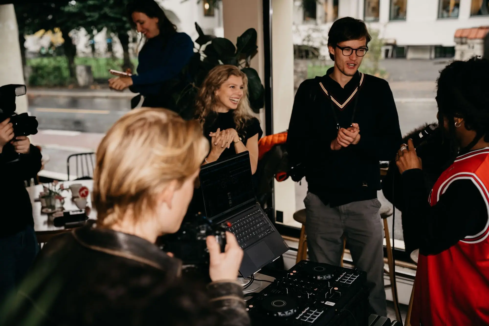 Guests gather around the DJ decks by the window while a man in a knit vest talks to the room.