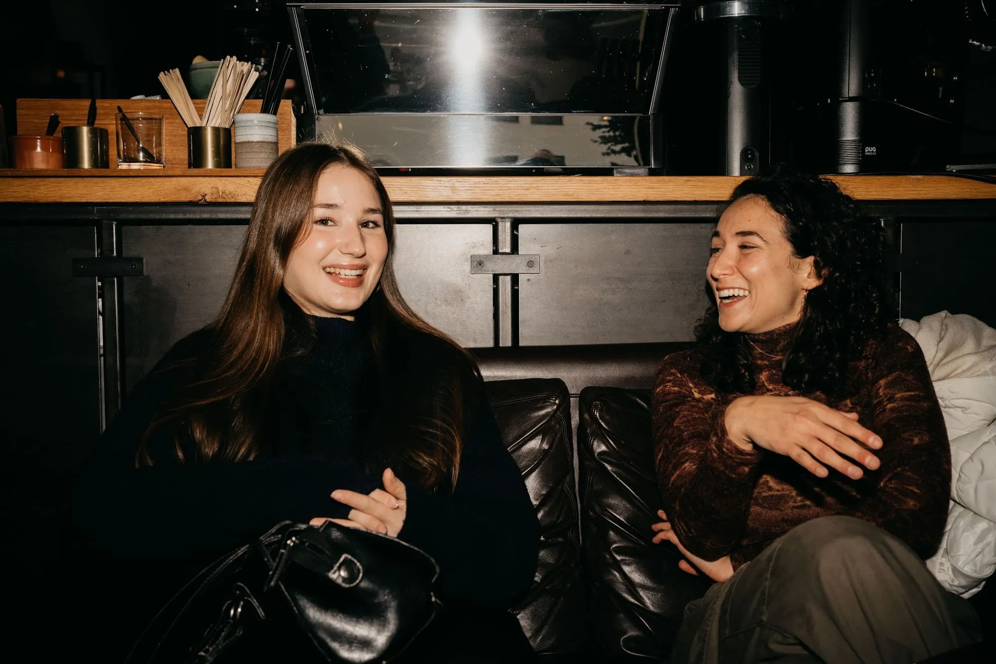 Two women laugh together on a leather sofa at the back of the bar.