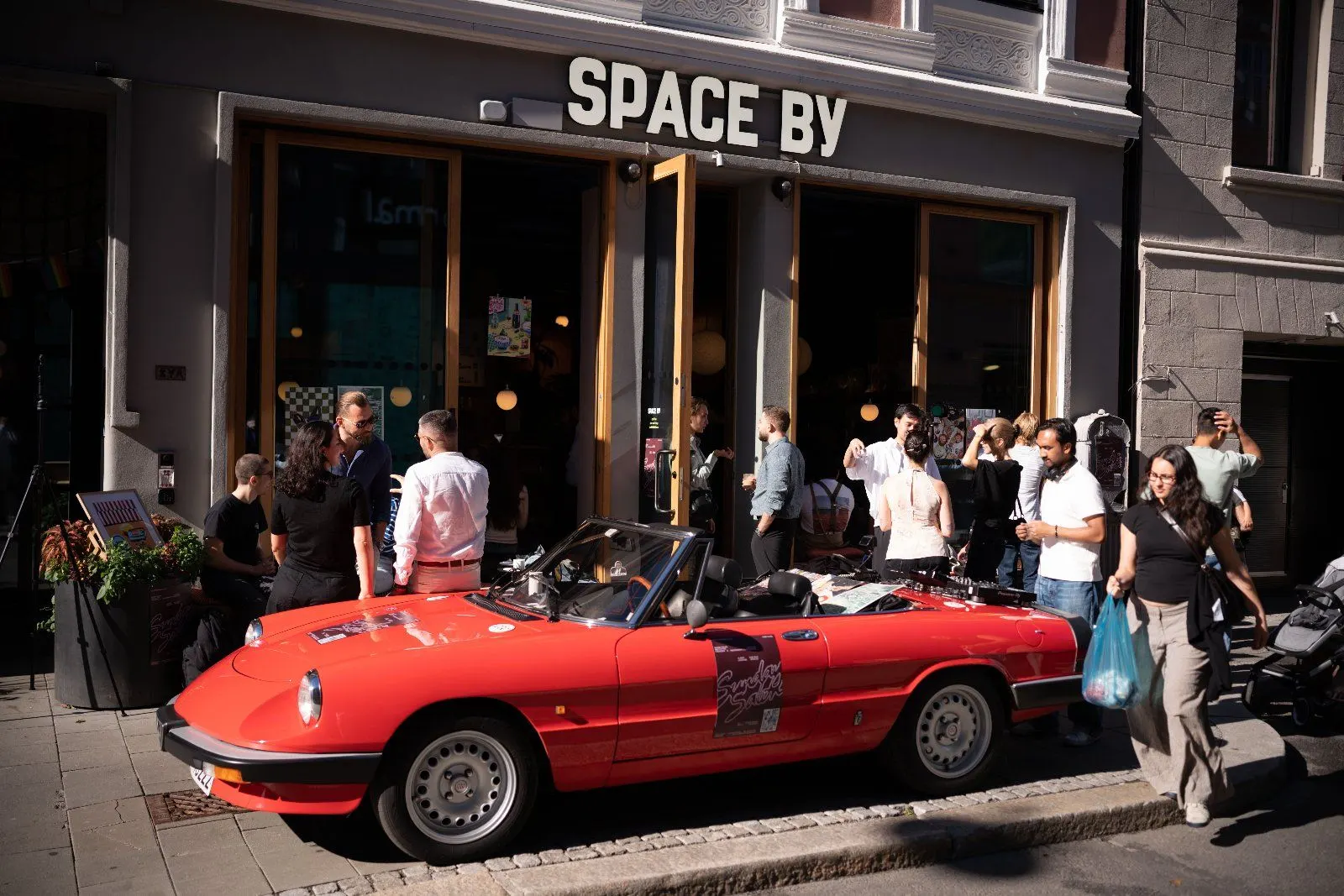The Space By storefront with the red convertible parked outside and a crowd of guests on the pavement.