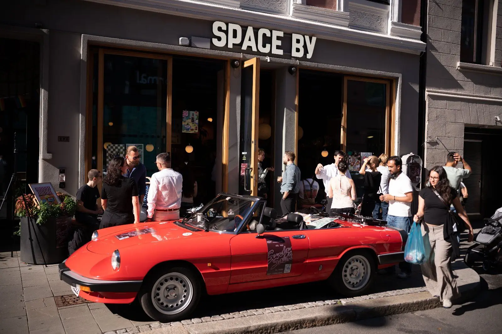 The Space By storefront with the red convertible parked outside and a crowd of guests on the pavement.