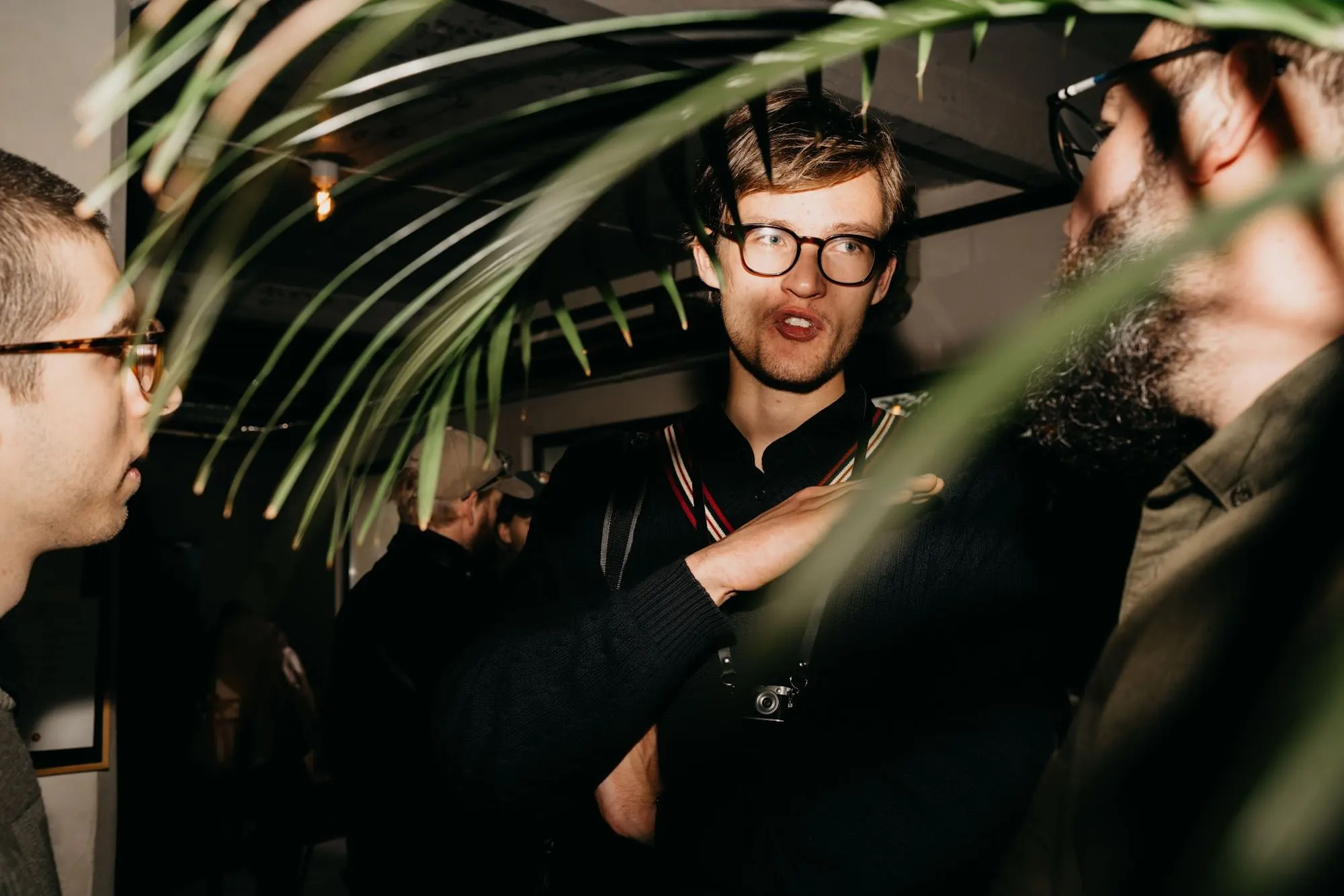 A man with glasses speaks to friends in a corner full of plants.