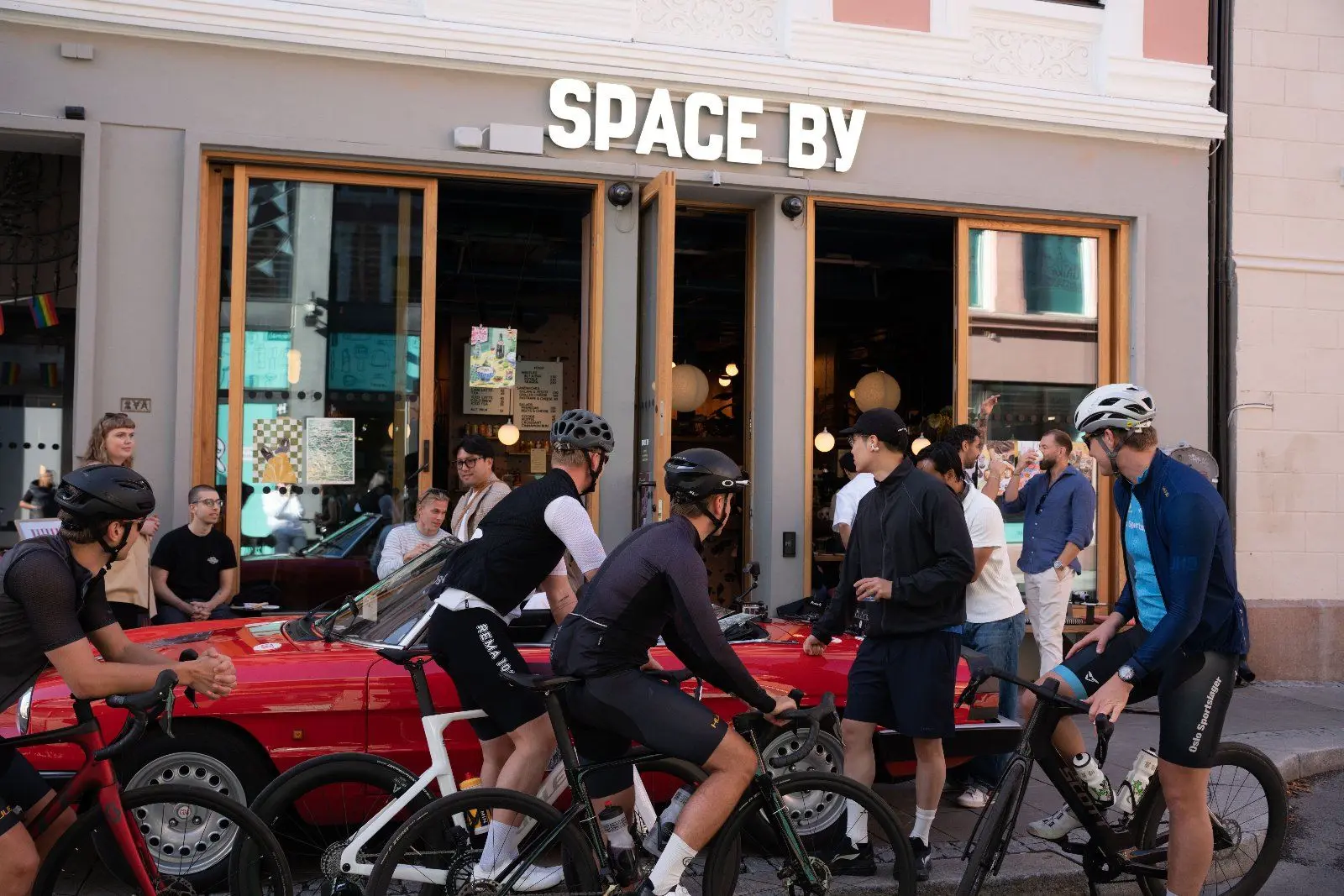 A group of cyclists in racing kit gather around the red car outside Space By.