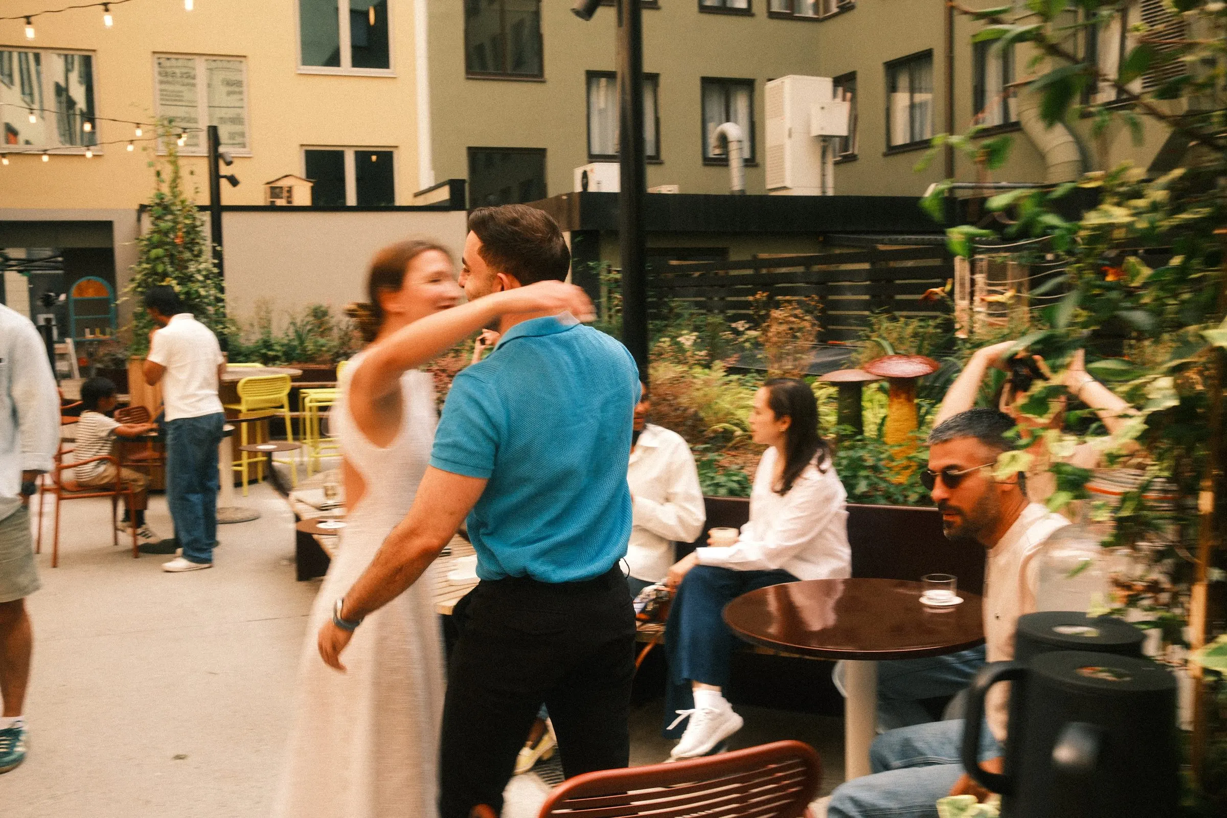 Two guests greet each other with a hug in a sunlit courtyard while others sit at café tables.