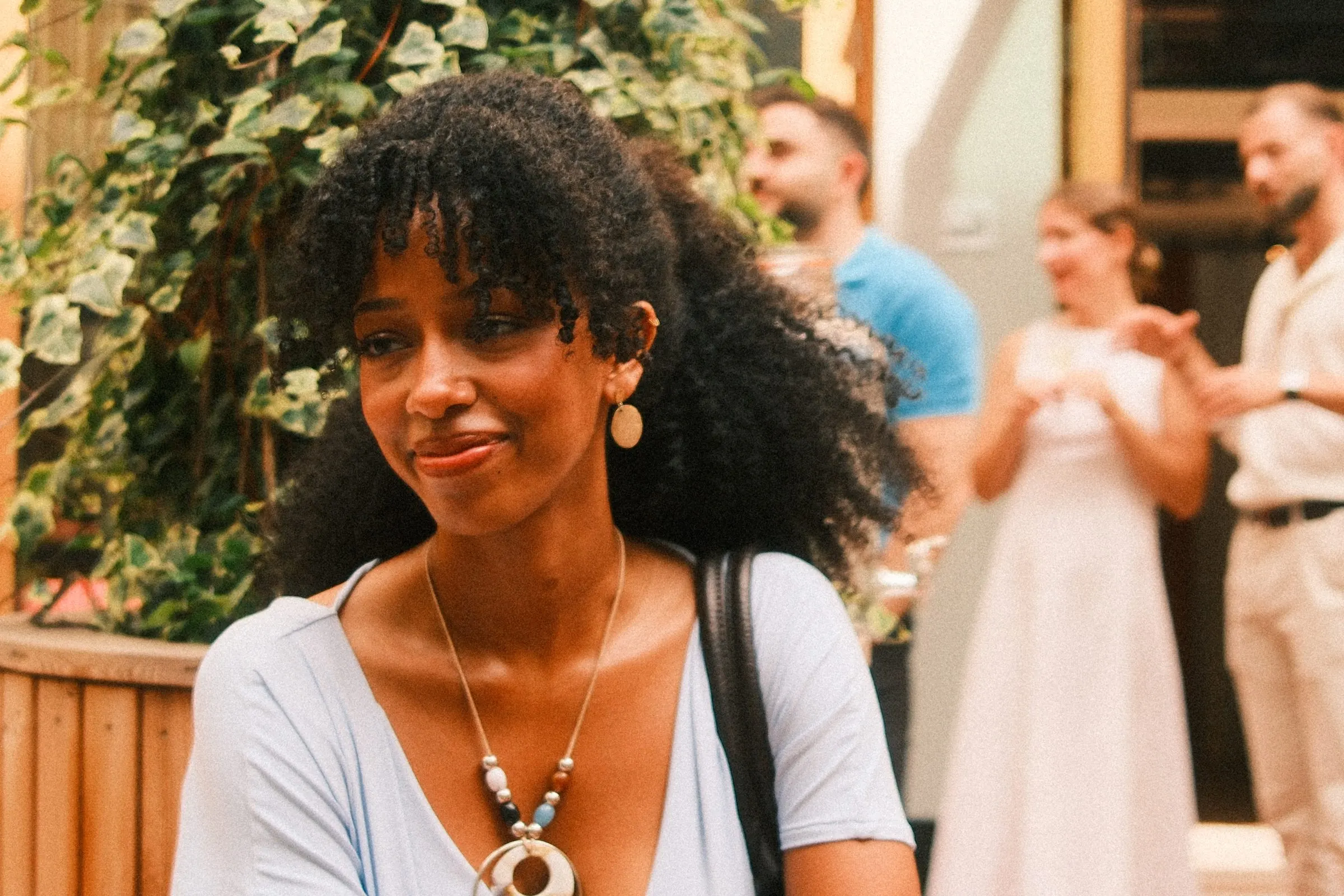 Close portrait of a smiling guest with curly hair and a beaded necklace, with the party blurred behind.