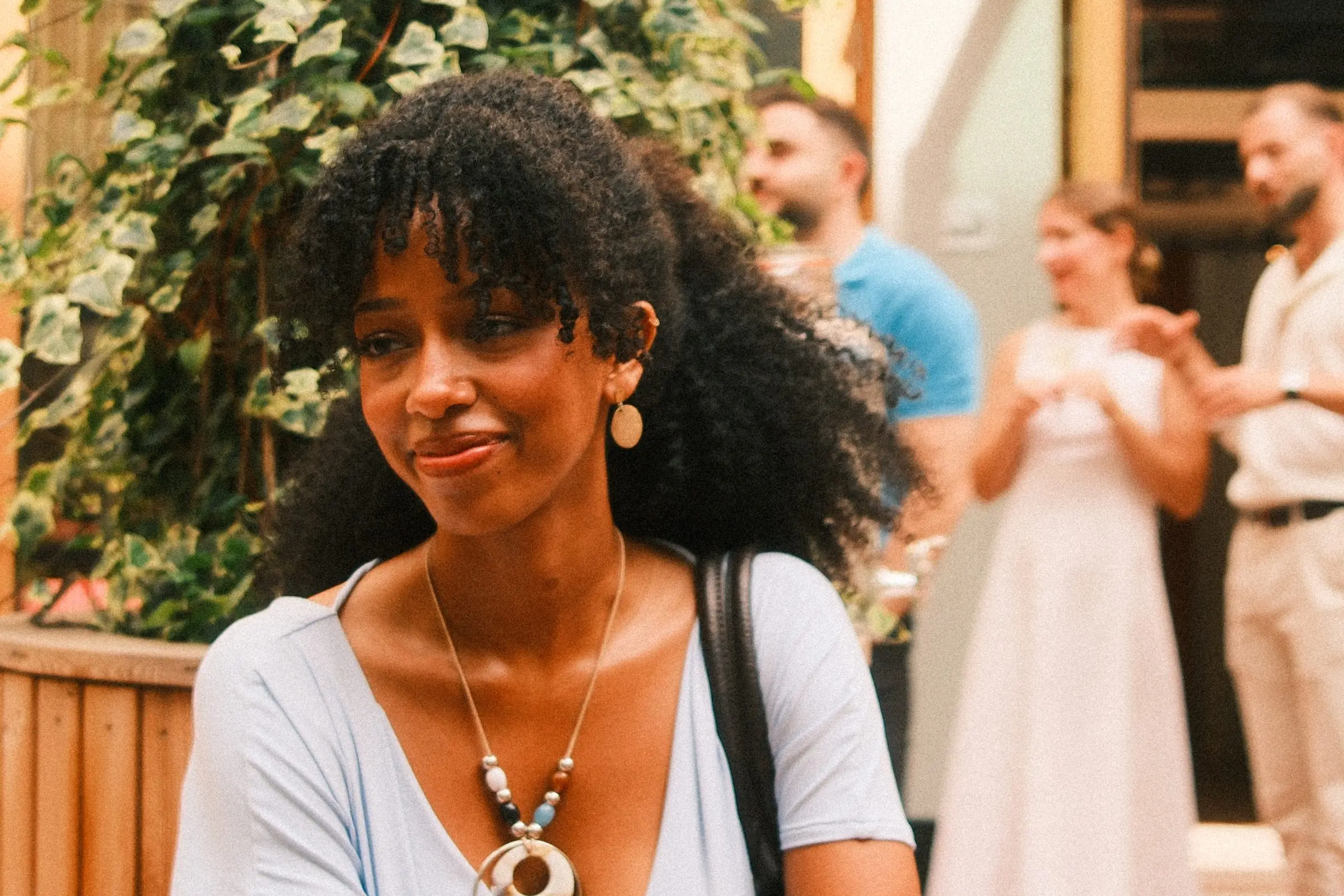 Close portrait of a smiling guest with curly hair and a beaded necklace, with the party blurred behind.