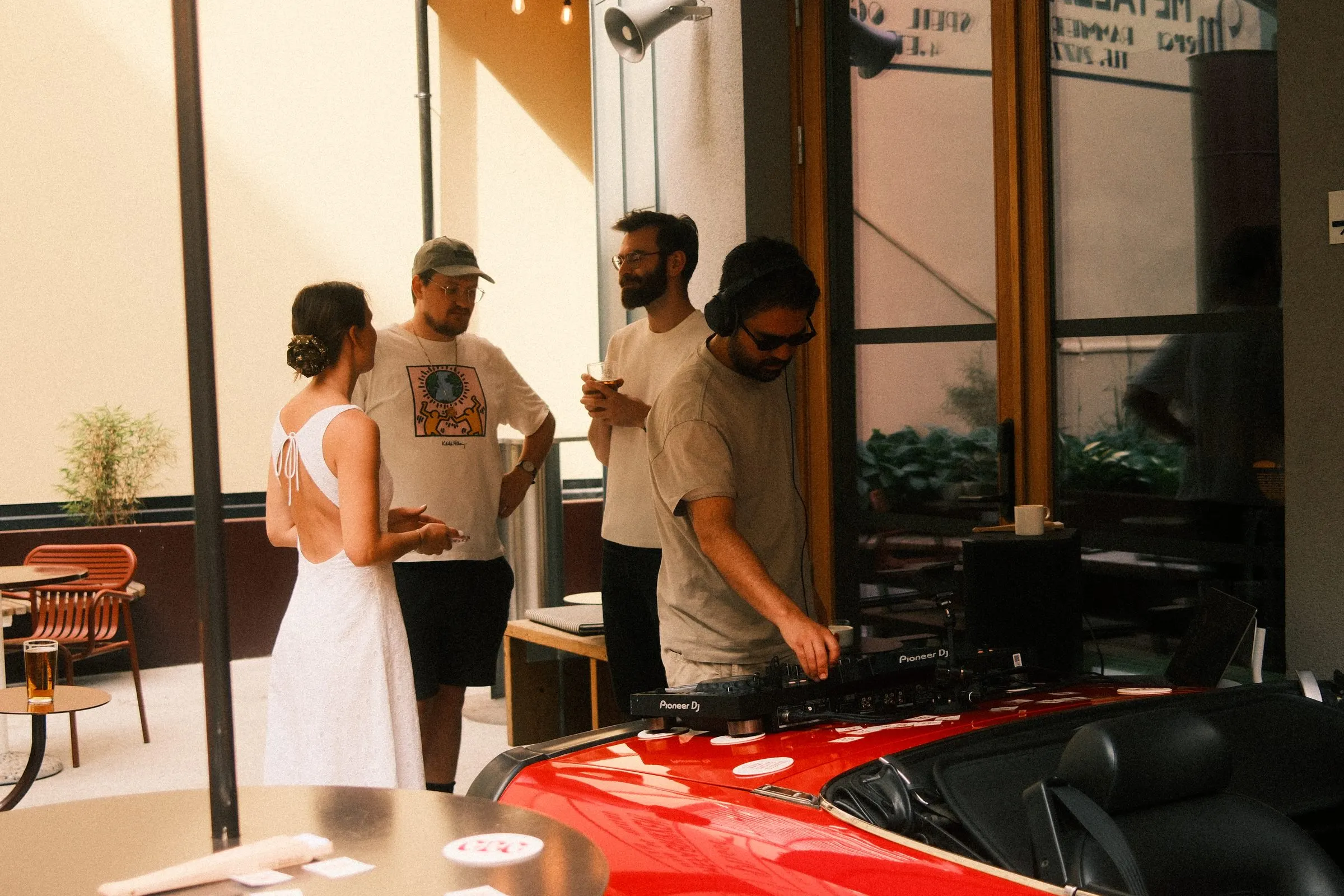 A DJ plays on decks set up on a red convertible while guests talk beside him.