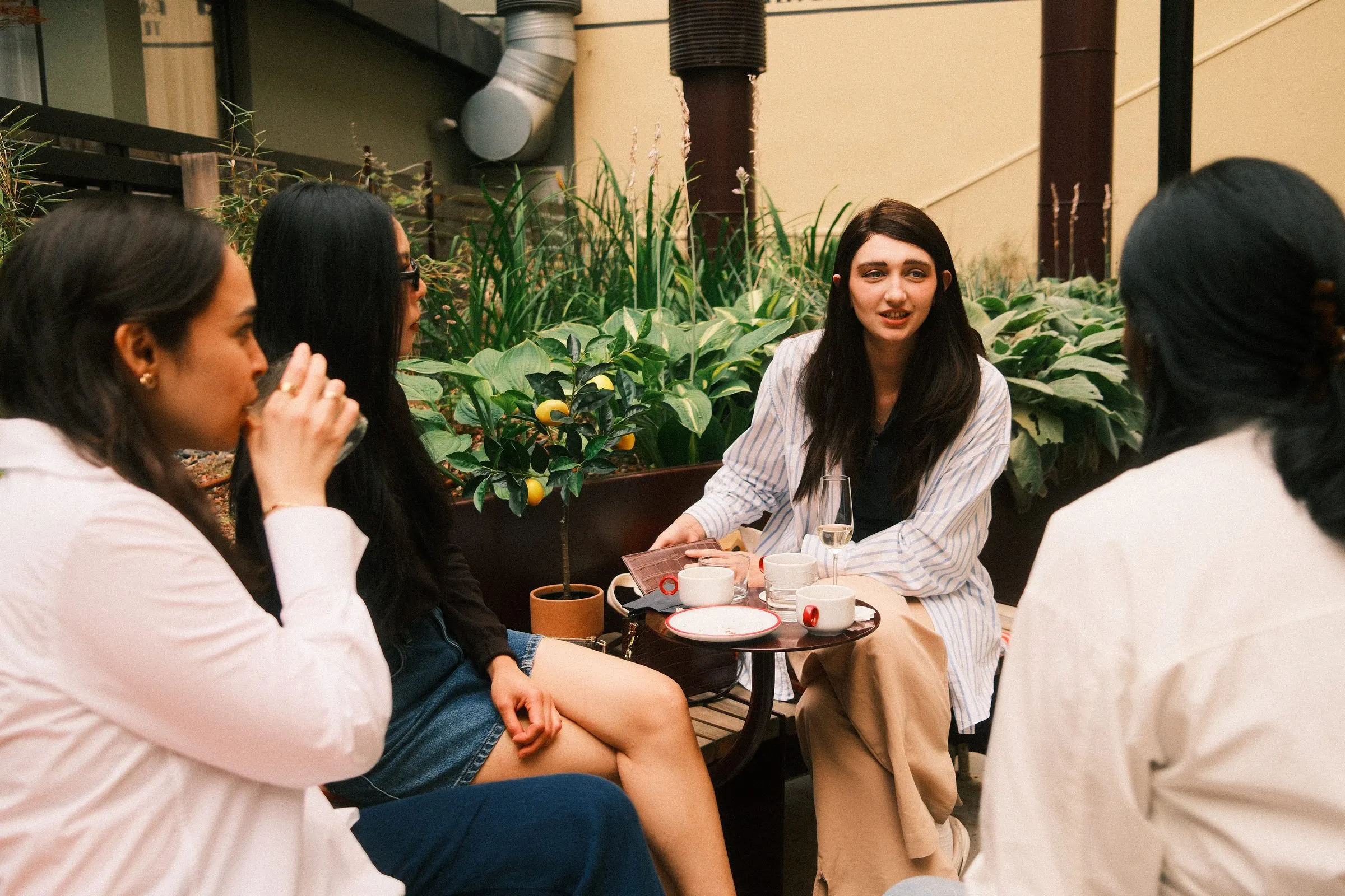 Guests sit around a small table with coffee cups among green plants, mid conversation.