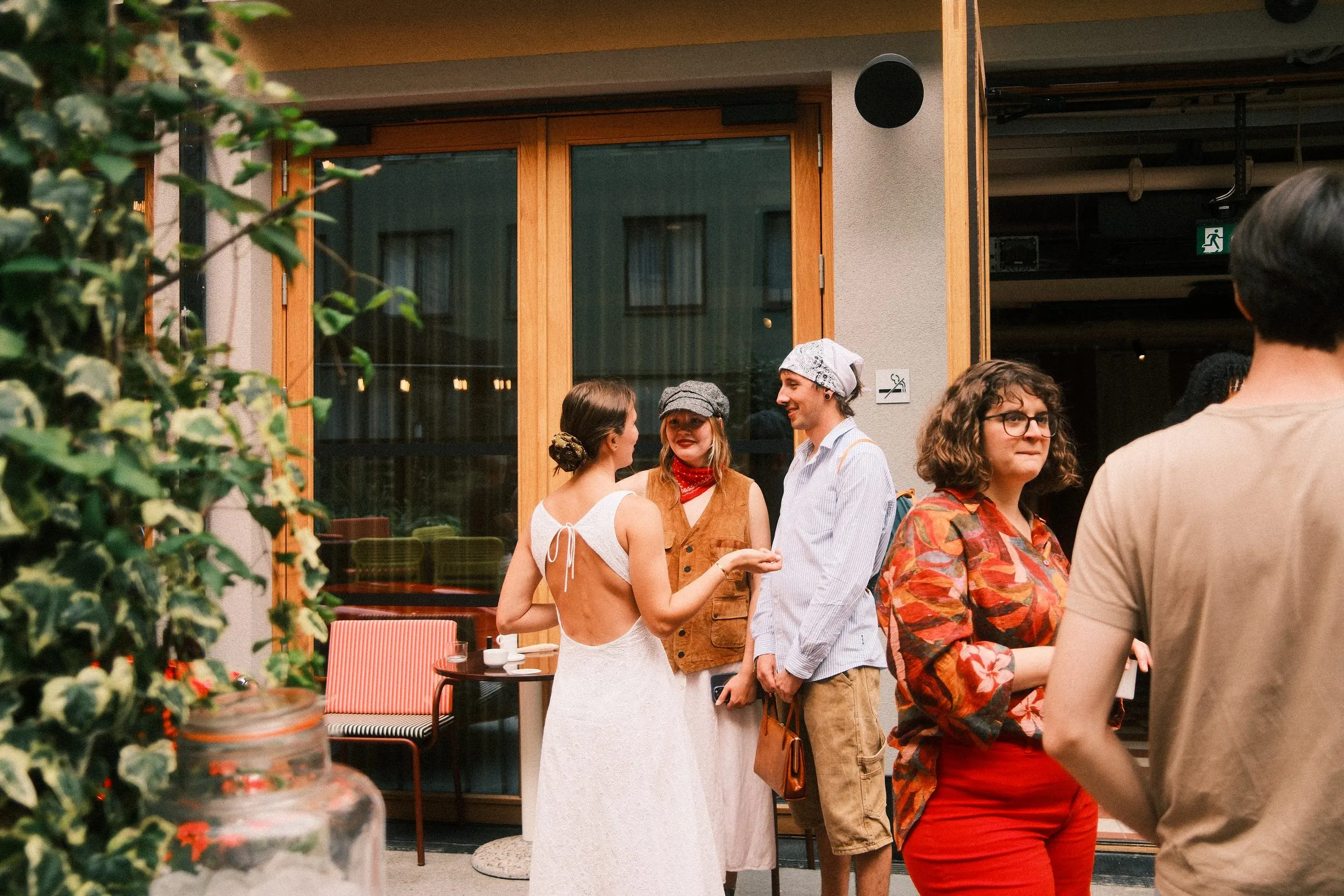 A group of guests chat outside the glass doors of the venue, framed by ivy and a table with coffee cups.