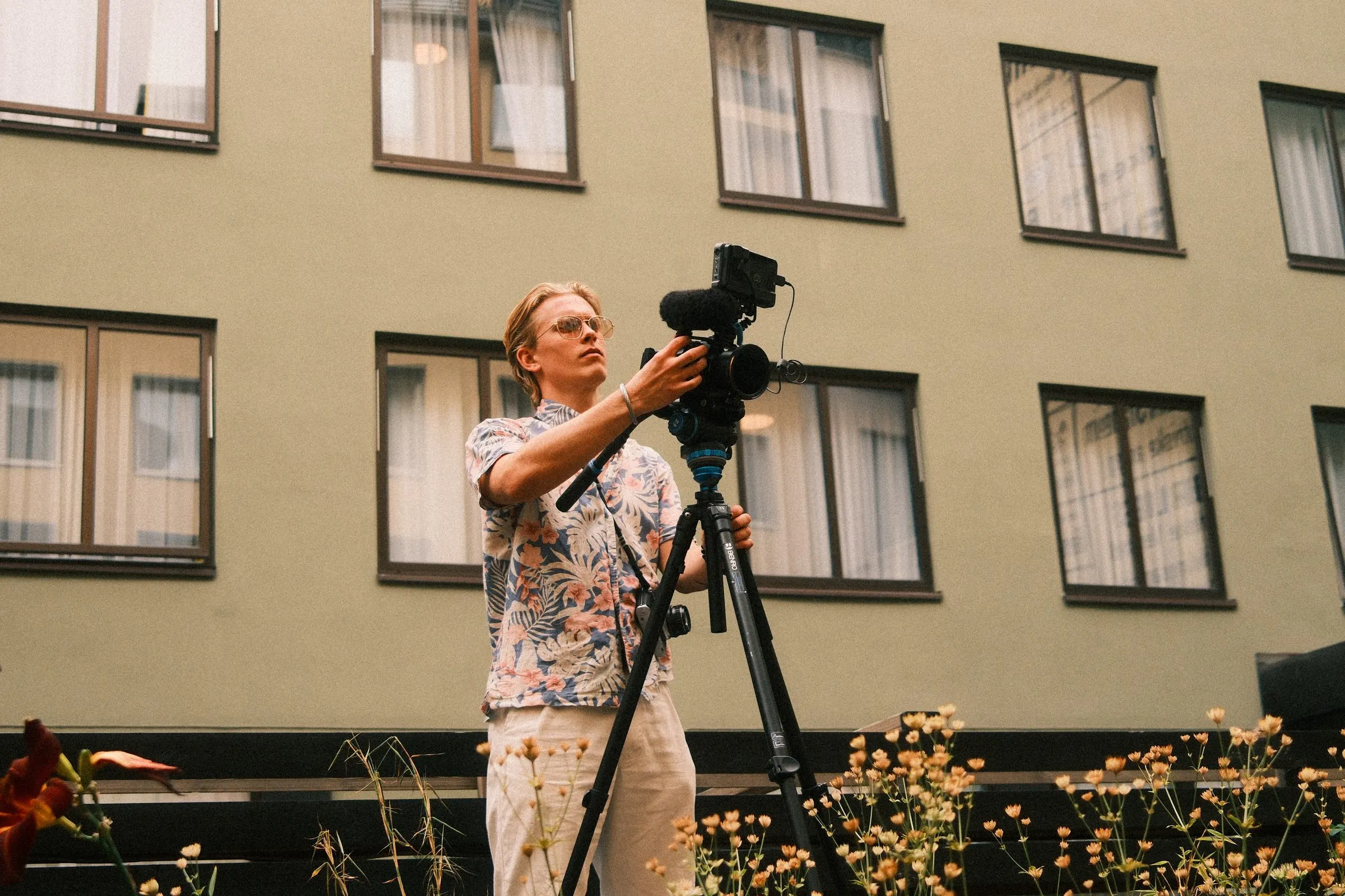A videographer in a floral shirt sets up a camera on a tripod in the courtyard.
