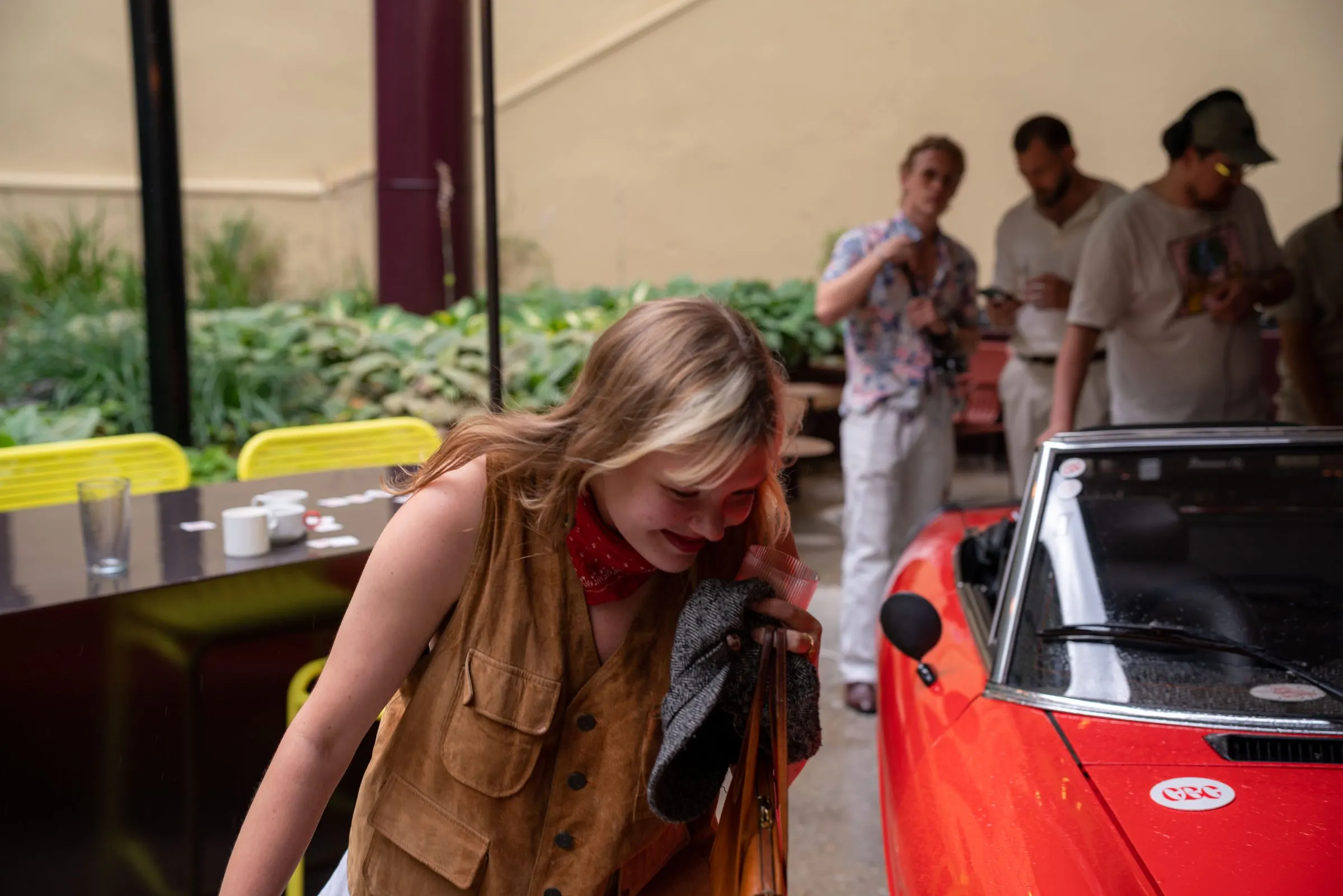 A guest in a suede vest laughs and looks down beside the red convertible while friends talk behind her.